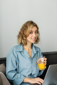 smiling young freelancer with wavy hair holding glass of orange juice near laptop