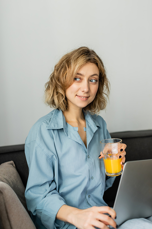 happy freelancer with wavy hair holding glass of orange juice near laptop