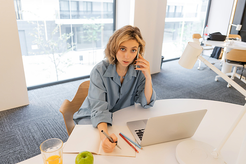 high angle view of young woman talking on smartphone near laptop on desk