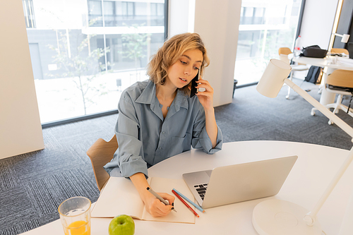young woman with wavy hair talking on smartphone near laptop on desk