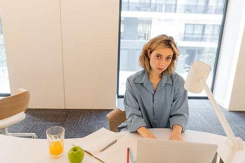 young woman with wavy hair using laptop near glass of orange juice and green apple on desk