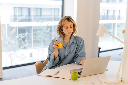 young woman with wavy hair holding glass of orange juice near laptop on desk