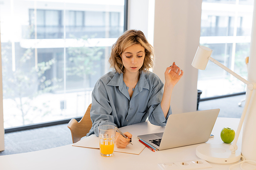 young woman with wavy hair writing in notebook near laptop on desk