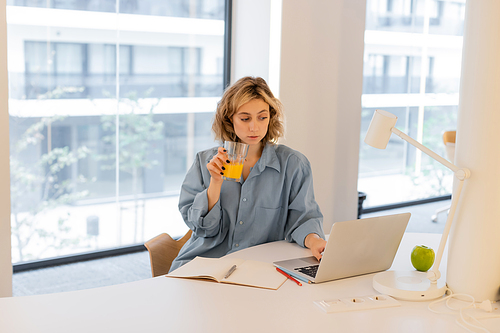 young woman with wavy hair holding glass of fresh orange juice while using laptop
