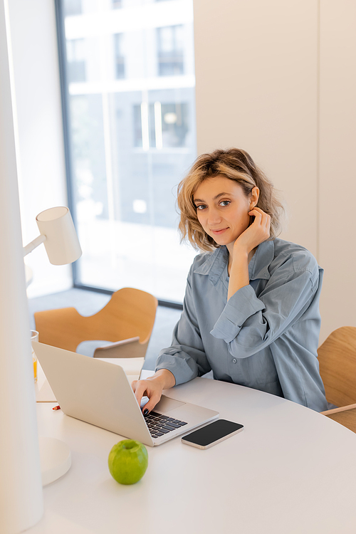 pleased young woman with wavy hair smiling while using laptop at home