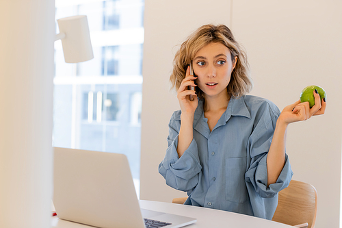 young woman with wavy hair talking on smartphone and holding green apple