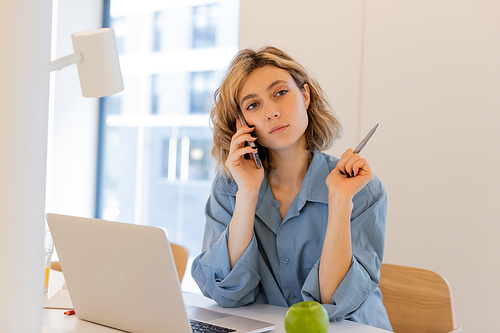 young woman with wavy hair talking on smartphone while holding pen