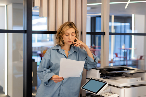 young blonde woman with wavy hair holding blank paper while standing near printer in office