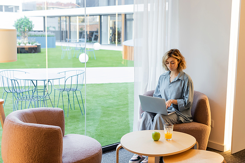 young blonde woman with wavy hair using laptop while sitting on armchair in lobby of hotel