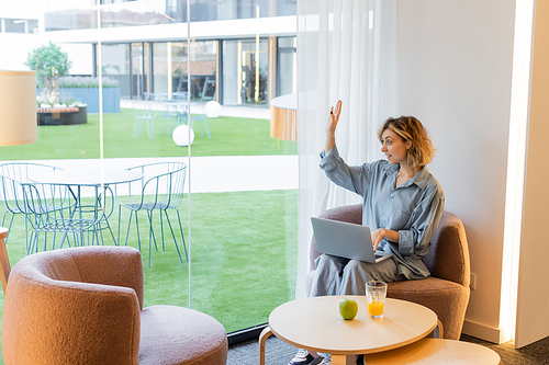 happy blonde woman with wavy hair waving hand while looking away and sitting with laptop in lobby of hotel