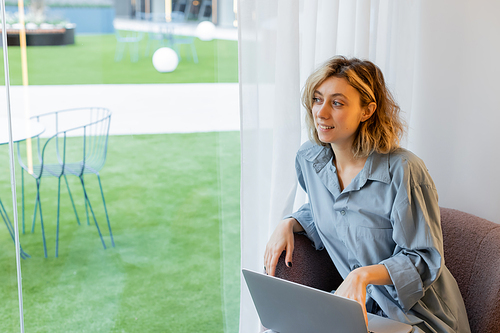 cheerful blonde woman with wavy hair looking away while sitting in armchair with laptop