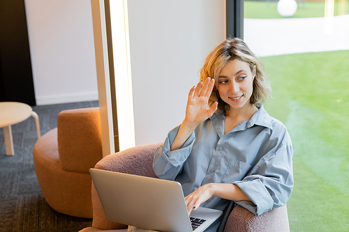happy blonde woman waving hand at window while looking away and sitting with laptop