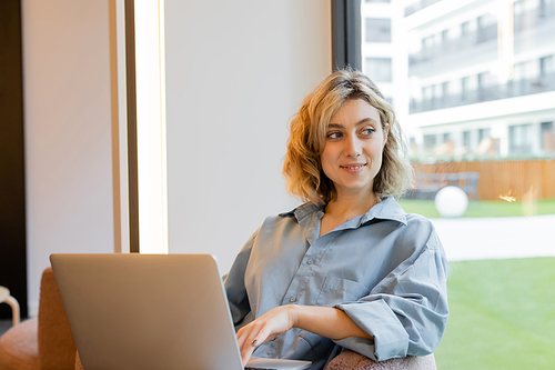 happy blonde woman with wavy hair looking at window and sitting with laptop