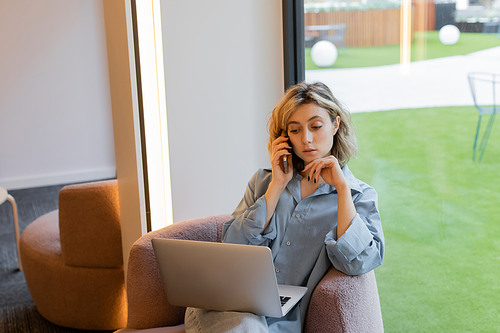 blonde freelancer with wavy hair talking on smartphone while using laptop in cafe