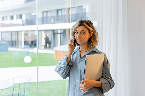 smiling young woman with wavy hair talking on smartphone and holding laptop near windows