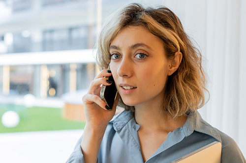 portrait of blonde young woman with wavy hair talking on smartphone near window