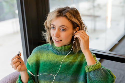 smiling young woman in green sweater wearing wired earphones while sitting in lobby of hotel in Barcelona