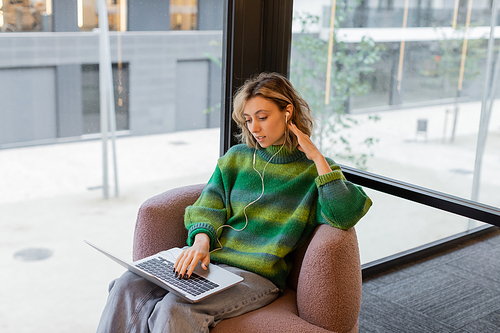 smiling young woman in sweater and wired earphones using laptop while sitting in lobby of hotel in Barcelona