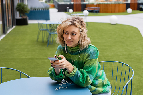 happy young woman in green sweater listening music in wired earphones and holding smartphone