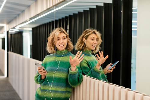 happy young woman listening music in wired earphones and holding smartphone while waving hand near mirrors in hotel