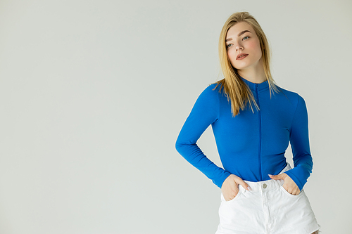 young woman in blue turtleneck standing with hands in pockets of white shorts isolated on grey
