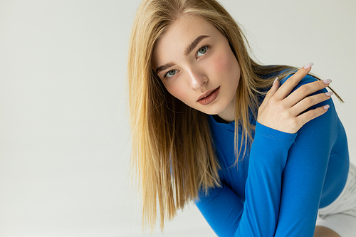 blonde woman with natural makeup wearing blue turtleneck and looking at camera isolated on grey