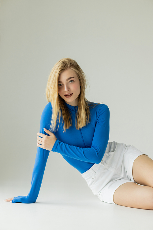 stylish blonde woman in blue turtleneck and white shorts smiling at camera while sitting on grey