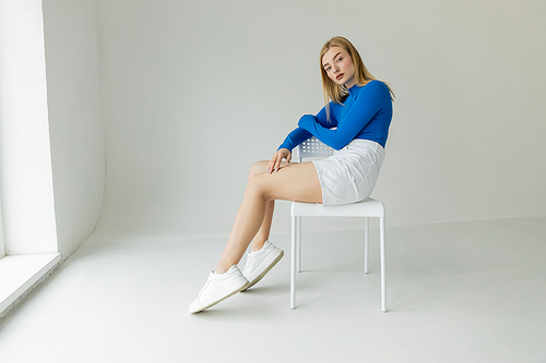 stylish woman in blue long sleeve shirt and white shorts sitting on chair and looking at camera on grey background