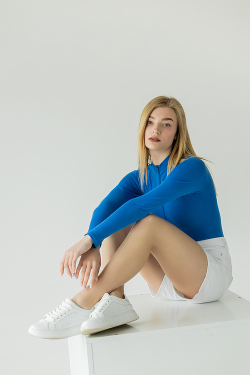 pretty woman in blue long sleeve shirt and sneakers sitting on white cube and looking at camera isolated on grey