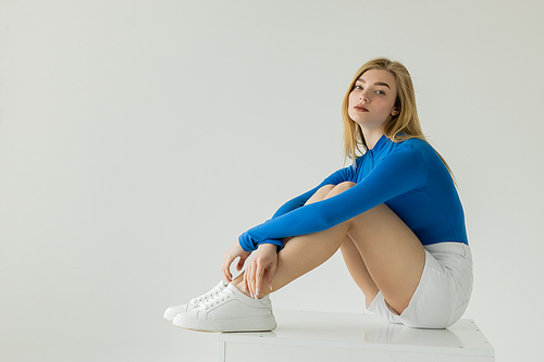 full length of blonde woman in blue long sleeve shirt and white sneakers sitting on cube and looking at camera isolated on grey