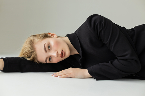 charming blonde woman in black blazer looking at camera while lying on white surface and grey background