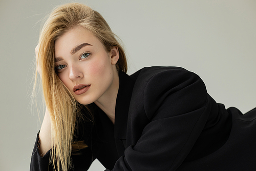 portrait of blonde woman with natural makeup wearing black jacket and looking at camera isolated on grey