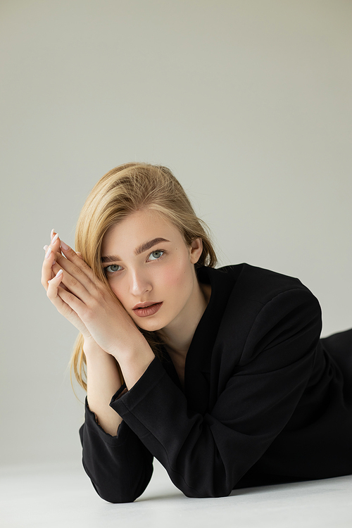 blonde woman with natural makeup lying in black jacket on white surface and grey background