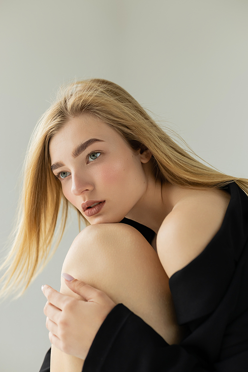 dreamy woman in black jacket leaning on knee and looking away isolated on grey