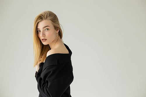 stylish blonde woman in black blazer posing with bare shoulder and looking at camera isolated on grey