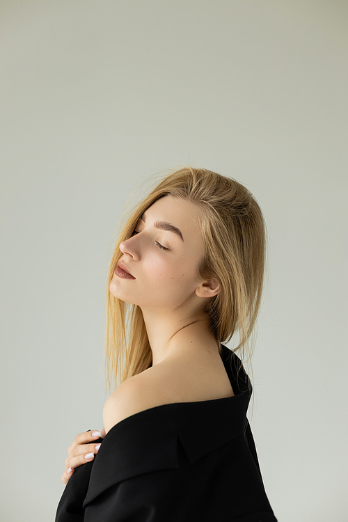 blonde and seductive woman in black blazer posing with closed eyes and bare shoulder isolated on grey