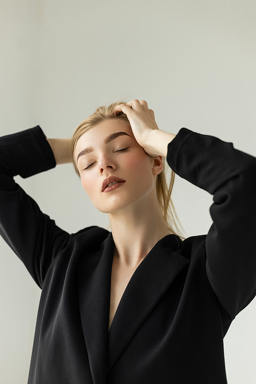 sensual young woman in black blazer touching blonde hair while posing with closed eyes isolated on grey