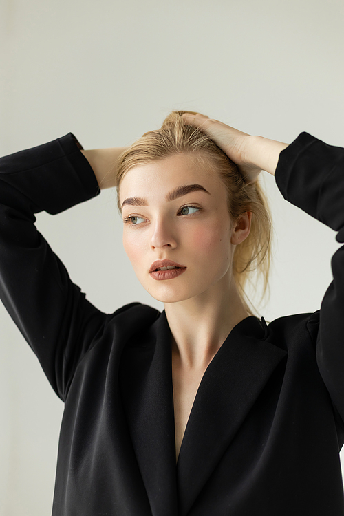 stylish blonde woman in black blazer adjusting hair and looking away isolated on grey