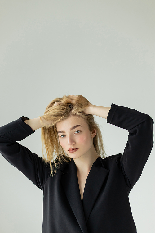 trendy woman in black blazer adjusting blonde hair and looking at camera isolated on grey