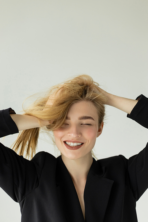 joyful woman in black jacket touching blonde hair and smiling with closed eyes isolated on grey