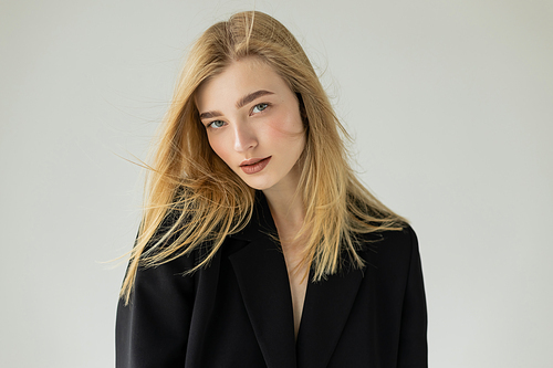 charming blonde woman with natural makeup wearing black blazer and looking at camera isolated on grey