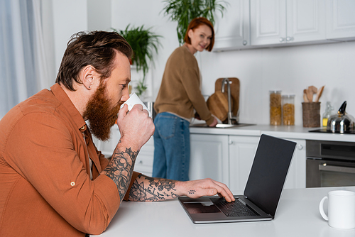 Bearded and tattooed man drinking coffee and using laptop near blurred wife in kitchen