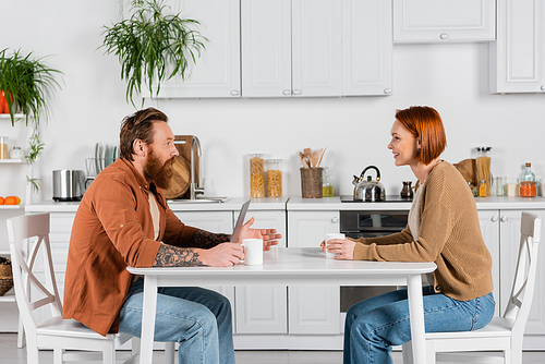 Side view of bearded man talking to redhead wife near laptop and cups in kitchen