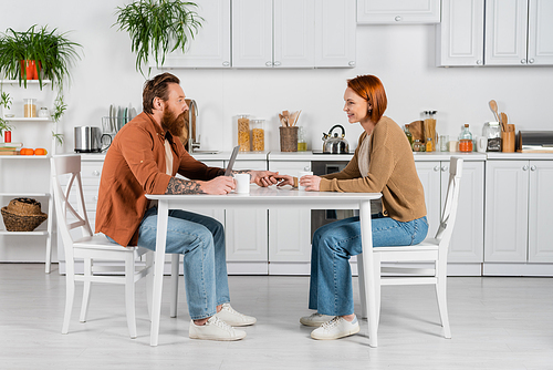 Side view of couple holding hands near laptop and cups in kitchen