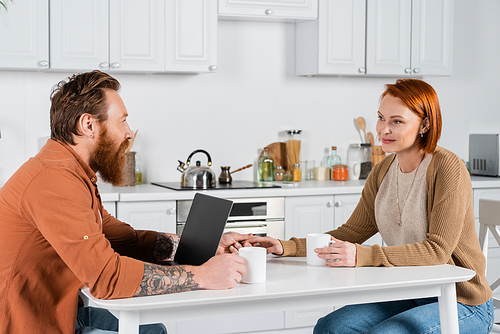 Adult couple holding hands near cups of coffee and laptop in kitchen