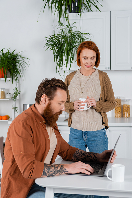 Redhead woman holding cup while husband using laptop in kitchen