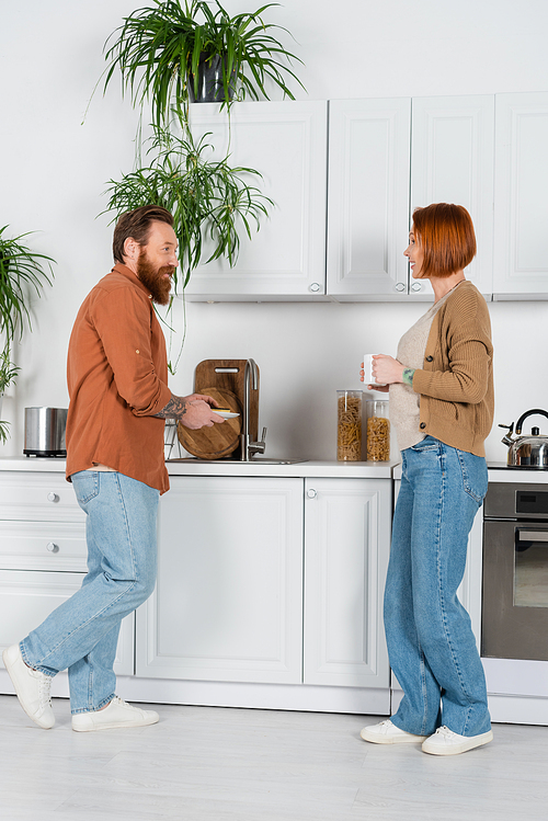 Side view of man washing plate near redhead wife with cup at home