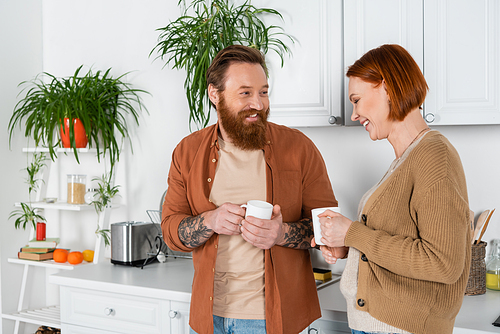 Smiling adult couple holding cups while talking in kitchen