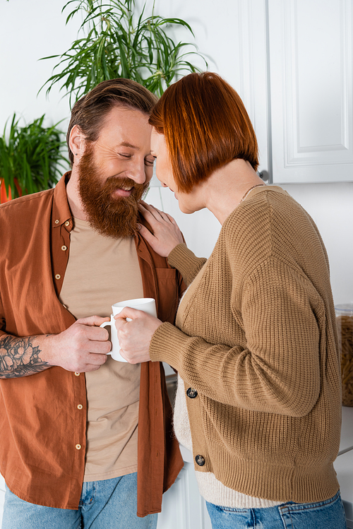 Redhead woman holding cup and touching cheerful bearded husband in kitchen