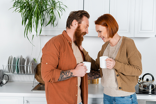 Side view of positive redhead woman holding cup and touching husband in kitchen